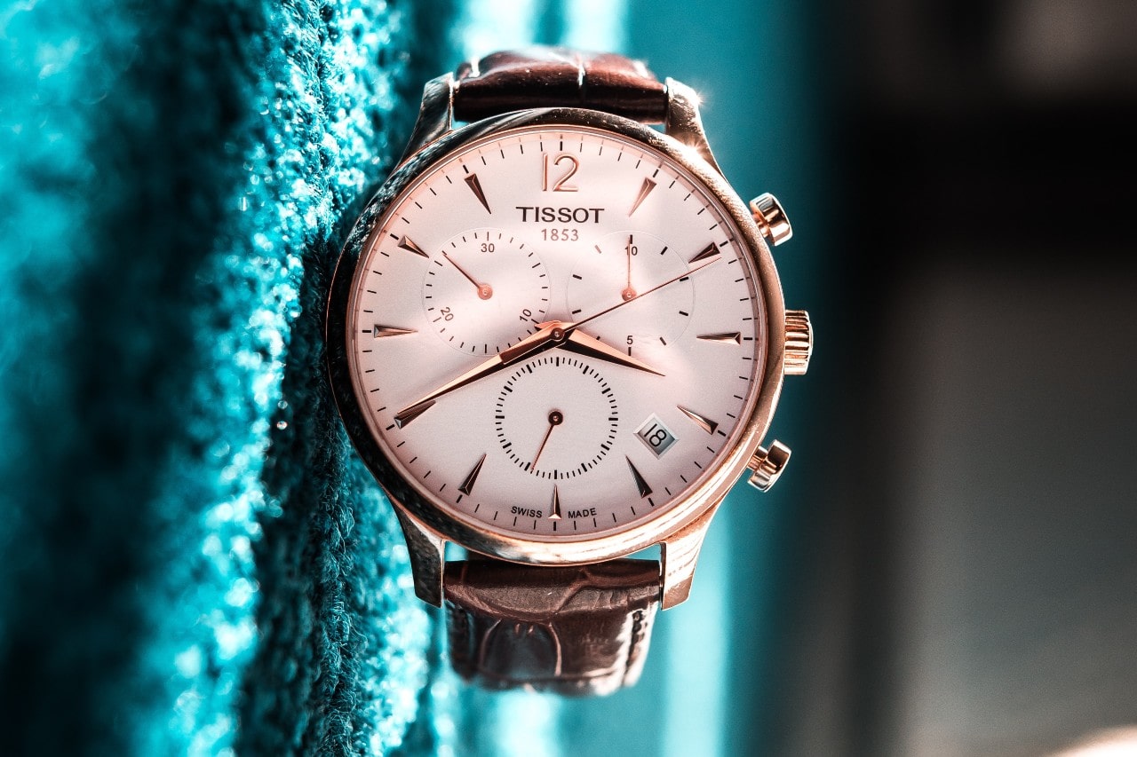 A rose gold watch case and brown alligator strap of a Tissot watch shown vertically while it’s laying on its side on a green carpet. A rose gold watch case and brown alligator strap of a Tissot watch shown vertically while it’s laying on its side on a green carpet.
