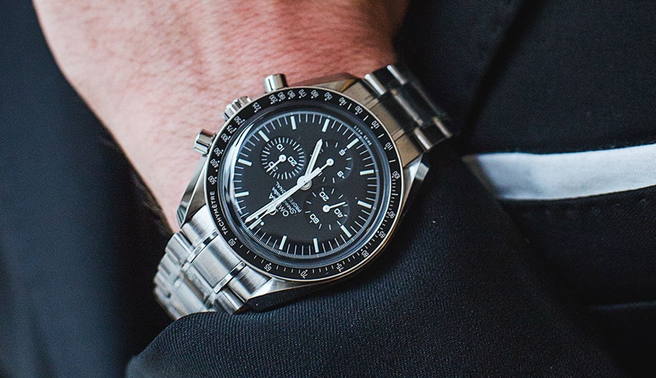 A close up of a man in a suit jacket’s Omega Speedmaster watch with a black watch dial and sterling silver bracelet along with multiple complications. A close up of a man in a suit jacket’s Omega Speedmaster watch with a black watch dial and sterling silver bracelet along with multiple complications.